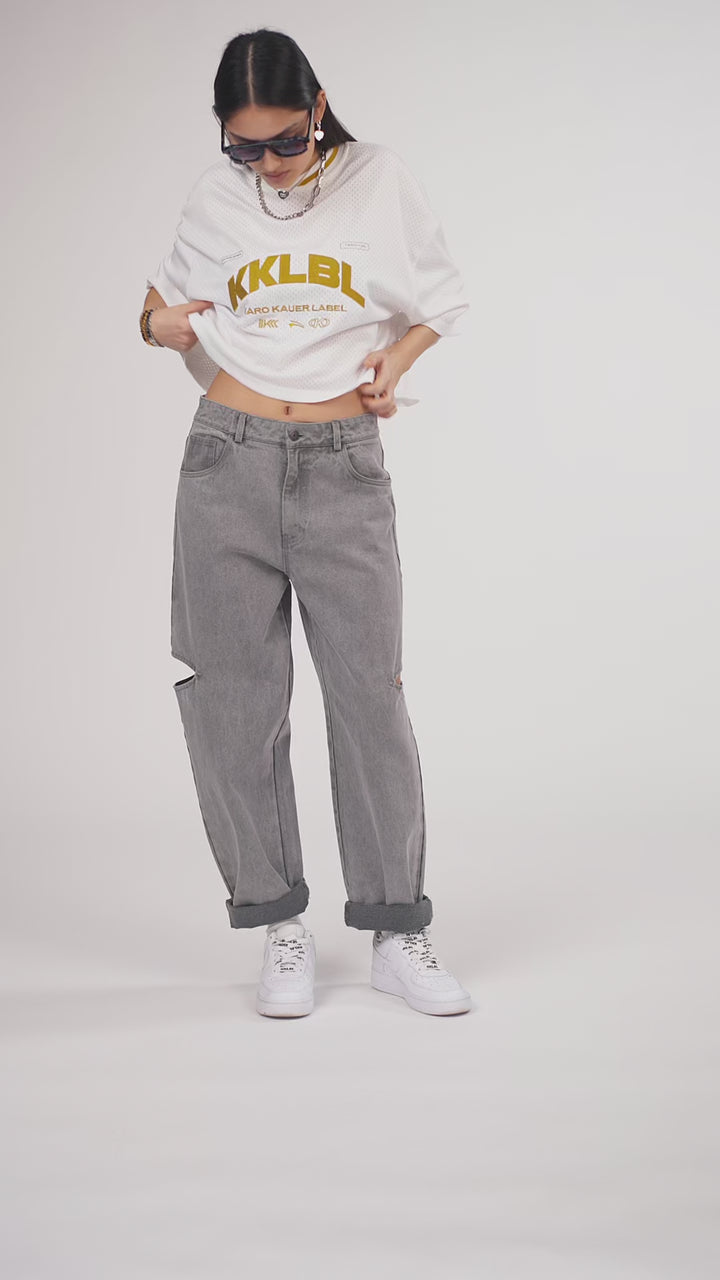 Ein Model in Ganzkörperansicht, das den Look in Bewegung aus verschiedenen Perspektiven zeigt, trägt ein weißes oversized Jersey-Shirt mit goldenem „KKLBL“-Print, kombiniert mit einer grauen Cut-out-Jeans aus der No Hustle Culture Kollektion, weißen Sneakern, Sonnenbrille und silberfarbenem Schmuck