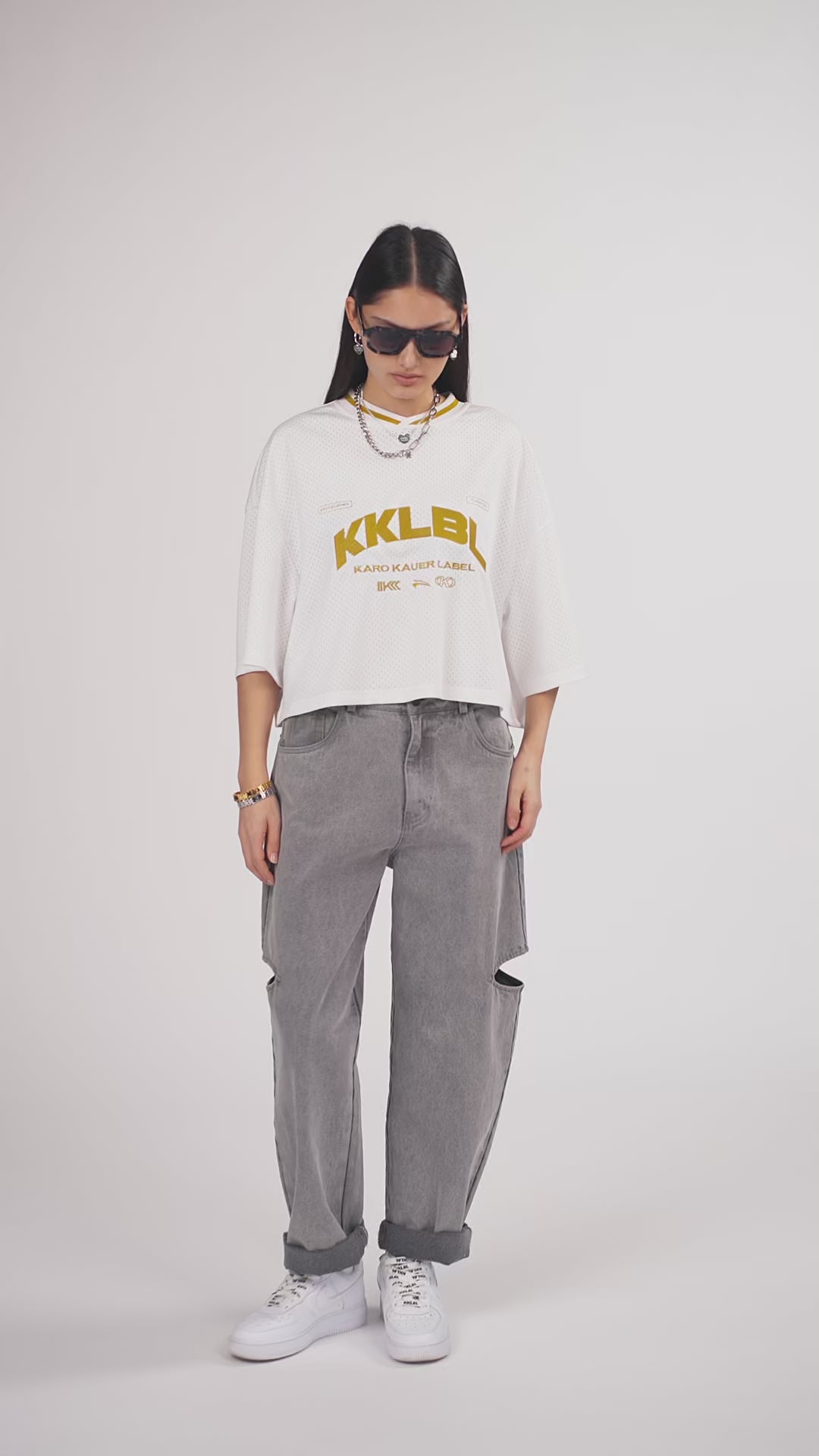 Ein Model in Ganzkörperansicht, das den Look in Bewegung aus verschiedenen Perspektiven zeigt, trägt ein weißes cropped Jersey mit großem goldenem „KKLBL“-Print auf der Brust, kombiniert mit einer grauen Cut-out-Jeans aus der No Hustle Culture Kollektion, weißen Sneakern, schwarzer Sonnenbrille und silberfarbenem Schmuck