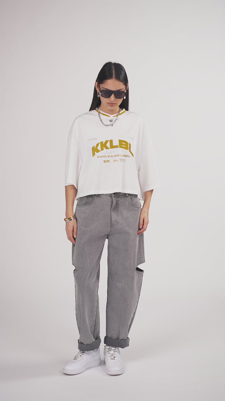 Ein Model in Ganzkörperansicht, das den Look in Bewegung aus verschiedenen Perspektiven zeigt, trägt ein weißes cropped Jersey mit großem goldenem „KKLBL“-Print auf der Brust, kombiniert mit einer grauen Cut-out-Jeans aus der No Hustle Culture Kollektion, weißen Sneakern, schwarzer Sonnenbrille und silberfarbenem Schmuck