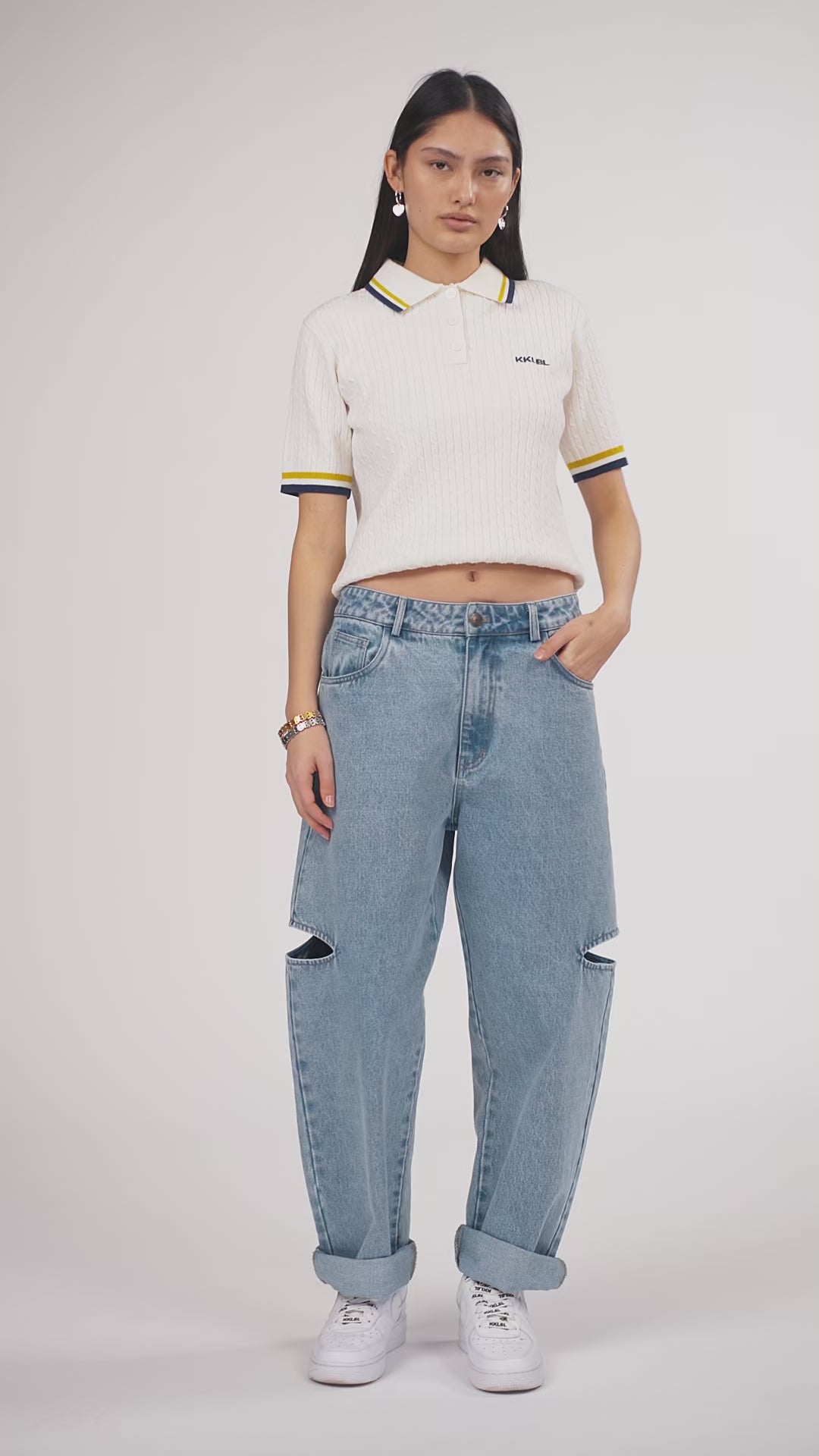 Ein Model in Ganzkörperansicht, das den Look in Bewegung zeigt, trägt ein hochgestecktes offwhite Strick-Poloshirt mit farblich abgesetztem Kragen, kleinem „KKLBL“-Schriftzug auf der Brust und kurzen Ärmeln, kombiniert mit einer hellblauen Cut-out-Jeans aus der No Hustle Culture Kollektion, weißen Sneakern und silberfarbenem Schmuck