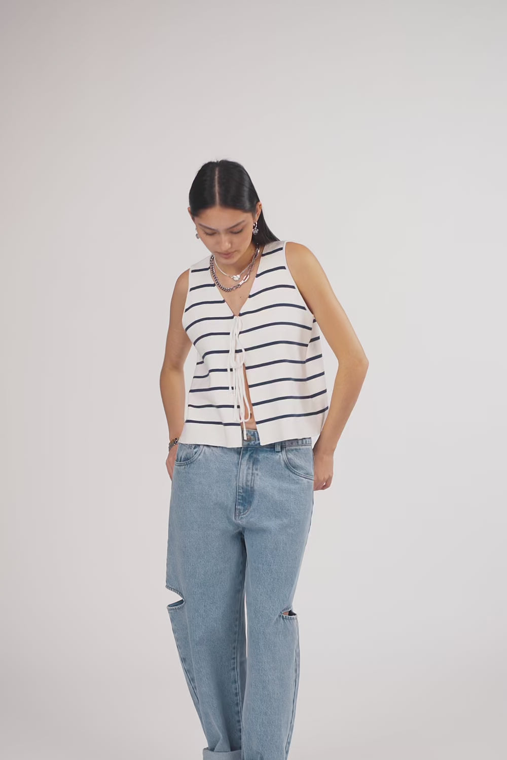 Ein Model in Ganzkörperansicht, das den Look in Bewegung zeigt, trägt eine ärmellose weiß-blau gestreifte gestrickte Weste mit Bindebändern vorn, kombiniert mit einer hellblauen Cut-out-Jeans aus der No Hustle Culture Kollektion und silberfarbenem Schmuck