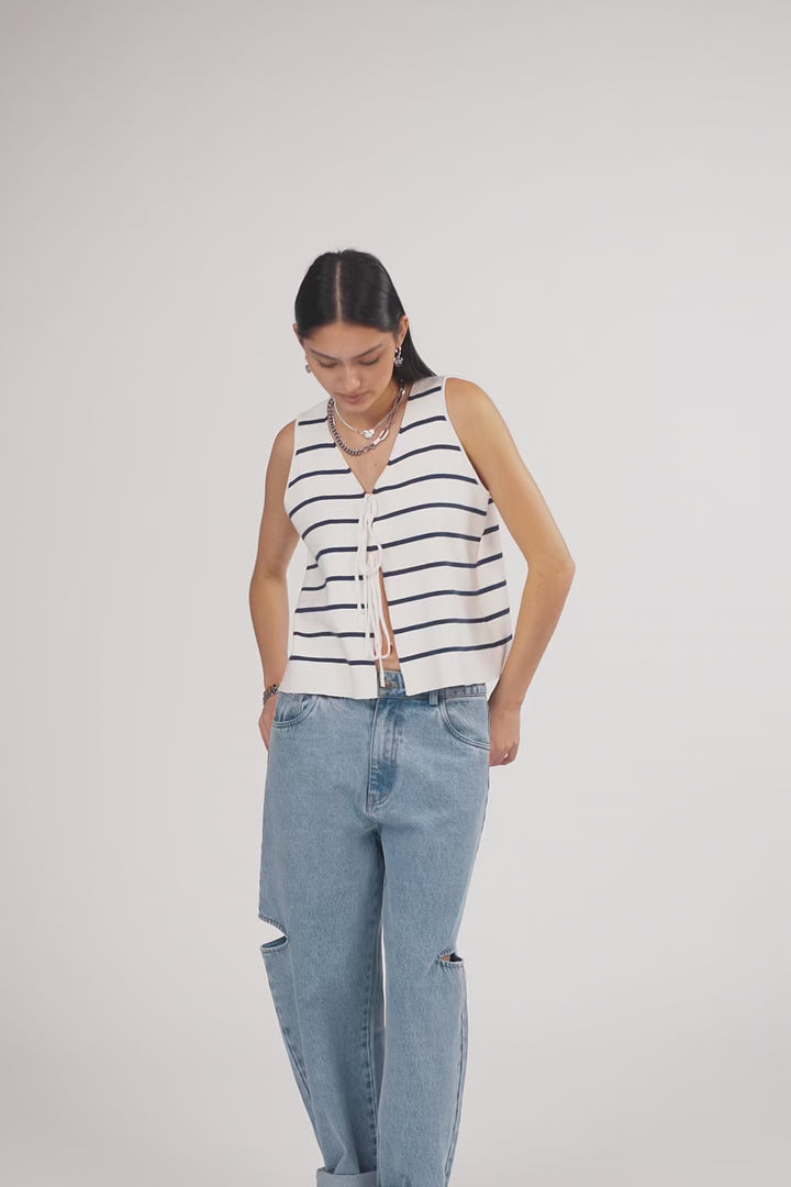 Ein Model in Ganzkörperansicht, das den Look in Bewegung zeigt, trägt eine ärmellose weiß-blau gestreifte gestrickte Weste mit Bindebändern vorn, kombiniert mit einer hellblauen Cut-out-Jeans aus der No Hustle Culture Kollektion und silberfarbenem Schmuck