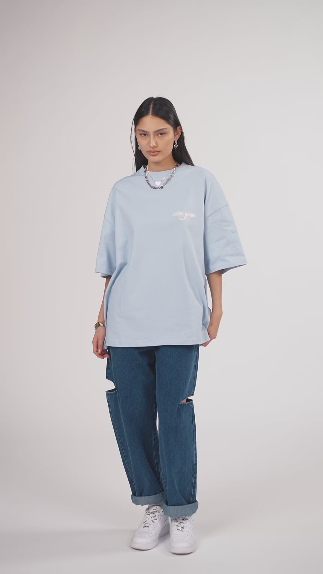 Ein Model in Ganzkörperansicht, das den Look in Bewegung aus verschiedenen Perspektiven zeigt, trägt ein hellblaues oversized T-Shirt mit mehreren weißen Prints, kombiniert mit einer blauen Cut-out-Jeans aus der No Hustle Culture Kollektion, weißen Sneakern und silberfarbenem Schmuck