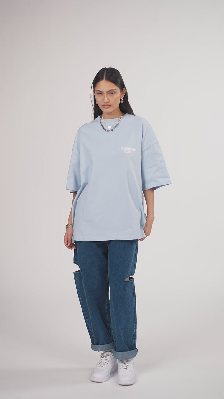 Ein Model in Ganzkörperansicht, das den Look in Bewegung aus verschiedenen Perspektiven zeigt, trägt ein hellblaues oversized T-Shirt mit mehreren weißen Prints, kombiniert mit einer blauen Cut-out-Jeans aus der No Hustle Culture Kollektion, weißen Sneakern und silberfarbenem Schmuck