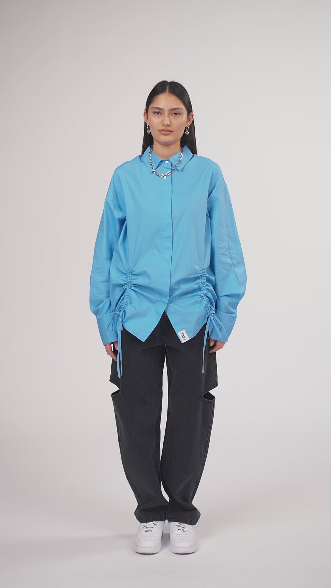 Ein Model in Ganzkörperansicht, das den Look in Bewegung aus verschiedenen Perspektiven zeigt, trägt eine hellblaue Bluse mit seitlichen Raffungen und Bindebändern, kombiniert mit einer schwarzen Cut-out-Jeans aus der No Hustle Culture Kollektion, weißen Sneakern und silberfarbenem Schmuck