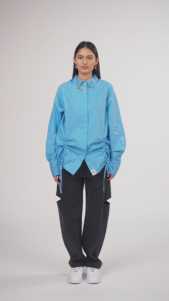 Ein Model in Ganzkörperansicht, das den Look in Bewegung aus verschiedenen Perspektiven zeigt, trägt eine hellblaue Bluse mit seitlichen Raffungen und Bindebändern, kombiniert mit einer schwarzen Cut-out-Jeans aus der No Hustle Culture Kollektion, weißen Sneakern und silberfarbenem Schmuck