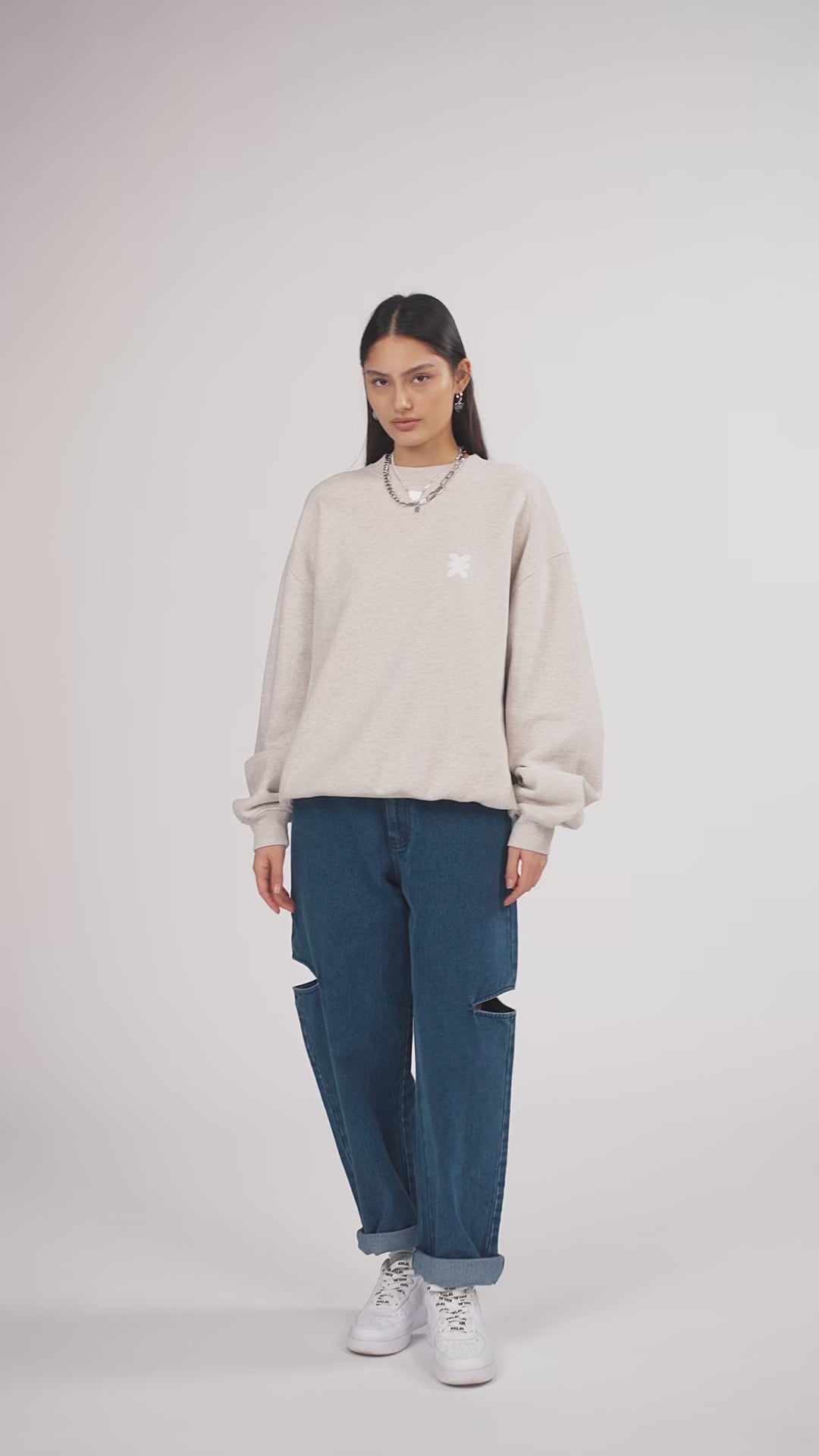 Ein Model in Ganzkörperansicht, das den Look in Bewegung aus verschiedenen Perspektiven zeigt, trägt ein hellgraues Oversize-Sweatshirt aus der No Hustle Culture Kollektion mit verschiedenen Prints, kombiniert mit einer blauen Cut-out-Jeans aus der No Hustle Culture Kollektion, weißen Sneakern und silberfarbenem Schmuck