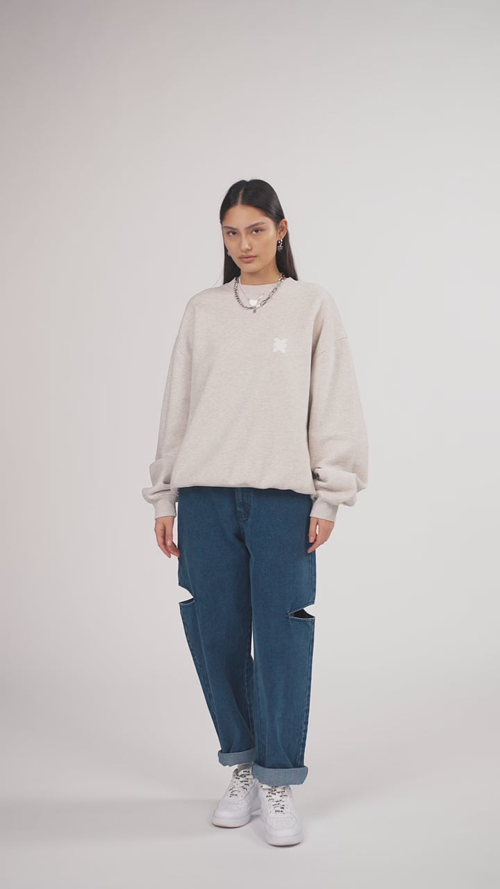 Ein Model in Ganzkörperansicht, das den Look in Bewegung aus verschiedenen Perspektiven zeigt, trägt ein hellgraues Oversize-Sweatshirt aus der No Hustle Culture Kollektion mit verschiedenen Prints, kombiniert mit einer blauen Cut-out-Jeans aus der No Hustle Culture Kollektion, weißen Sneakern und silberfarbenem Schmuck