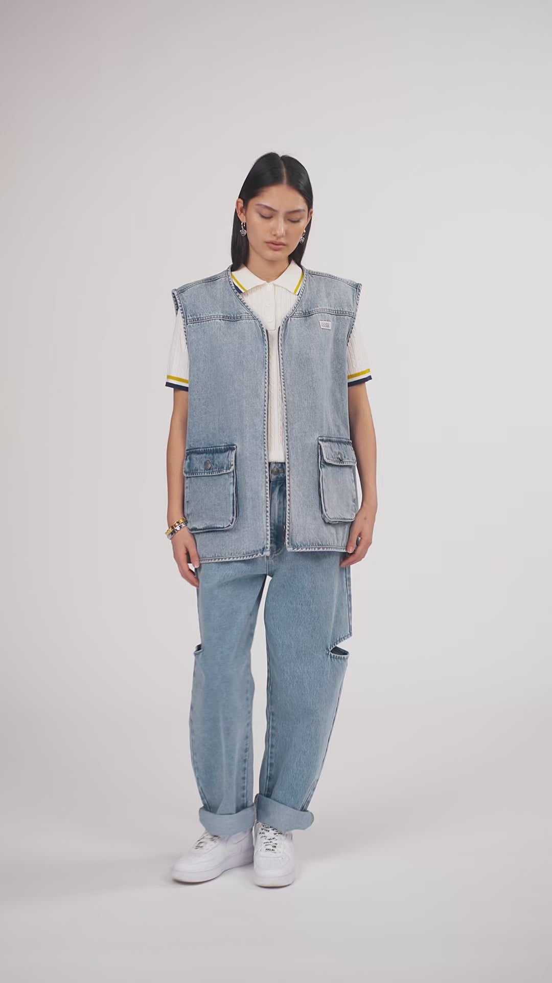Ein Model in Ganzkörperansicht, das den Look in Bewegung aus verschiedenen Perspektiven zeigt, trägt eine blaue Denimweste und eine blaue Cut-out-Jeans aus der No Hustle Culture Kollektion, kombiniert mit einem offwhite Strick-Poloshirt, weißen Sneakern und silberfarbenem Schmuck