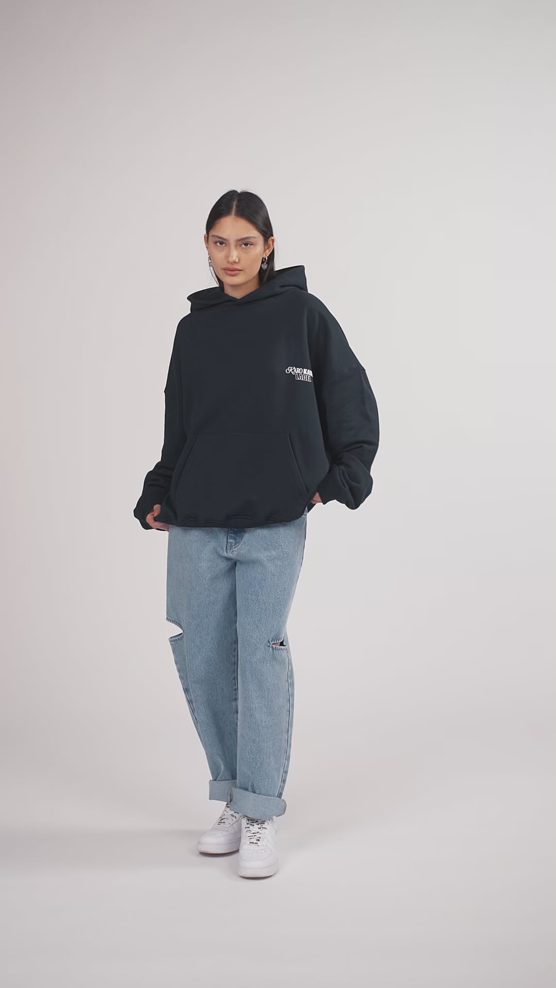 Ein Model in Ganzkörperansicht, das den Look in Bewegung aus verschiedenen Perspektiven zeigt und dabei die Kapuze aufsetzt, trägt einen dunkelblauen Oversize-Hoodie aus der No Hustle Culture Kollektion mit kleinem weißen Brustprint sowie einem größeren Rückenprint, kombiniert mit einer hellblauen Cut-out-Jeans aus der No Hustle Culture Kollektion und weißen Sneakern
