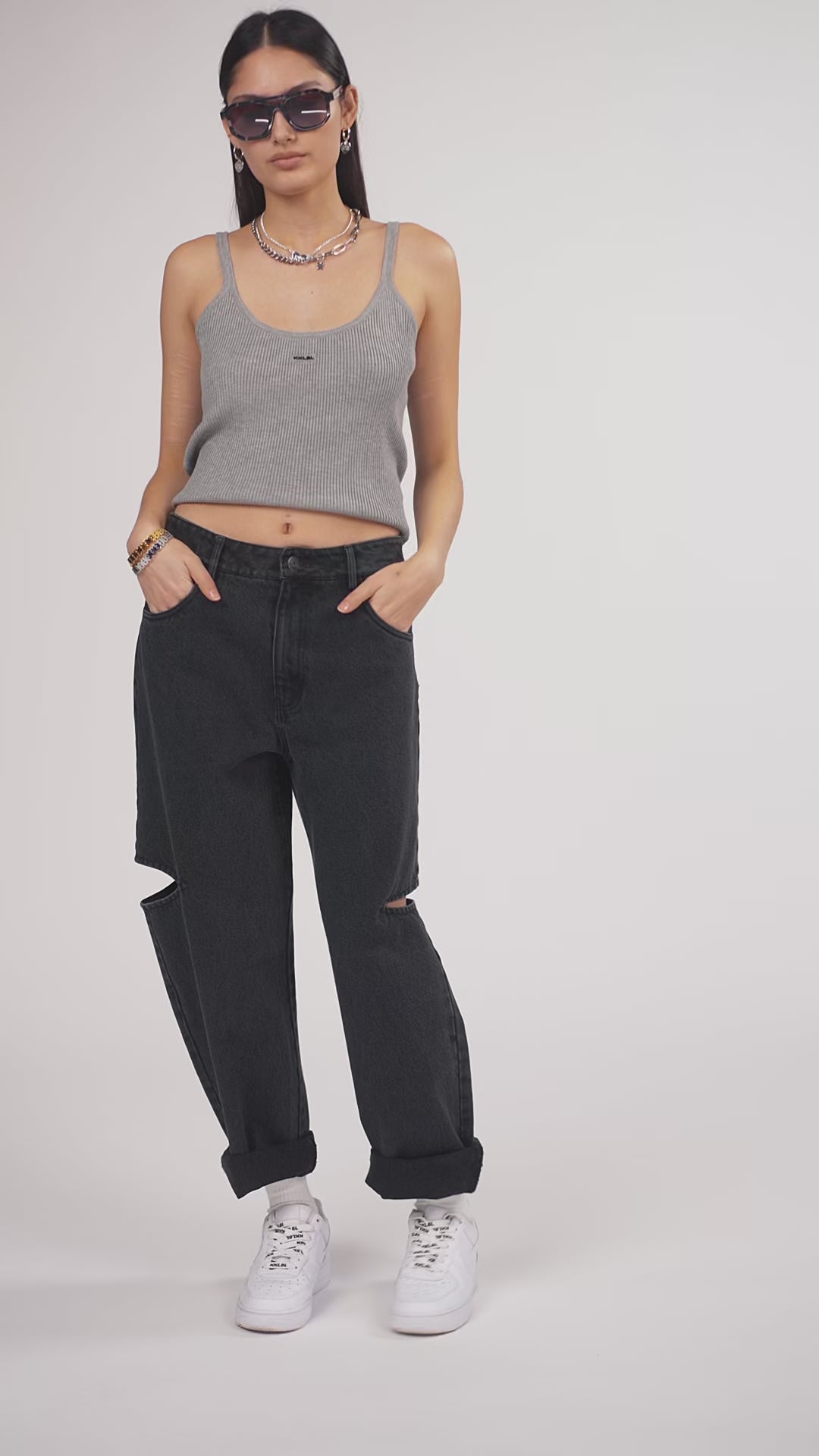 Ein Model in Ganzkörperansicht, das den Look in Bewegung aus verschiedenen Perspektiven zeigt, trägt eine schwarze Cut-out-Jeans und ein graues cropped Tanktop aus der No Hustle Kollektion, kombiniert mit weißen Sneakern, Sonnenbrille und silberfarbenem Schmuck