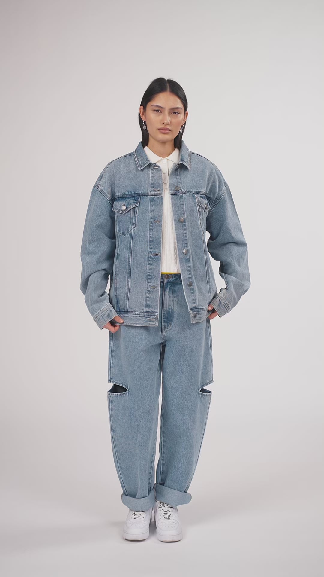 Ein Model in Ganzkörperansicht, das den Look in Bewegung aus verschiedenen Perspektiven zeigt, trägt eine blaue Denimjacke und eine blaue Cut-out-Jeans aus der No Hustle Culture Kollektion, kombiniert mit einem offwhite Strick-Poloshirt, weißen Sneakern und silberfarbenem Schmuck