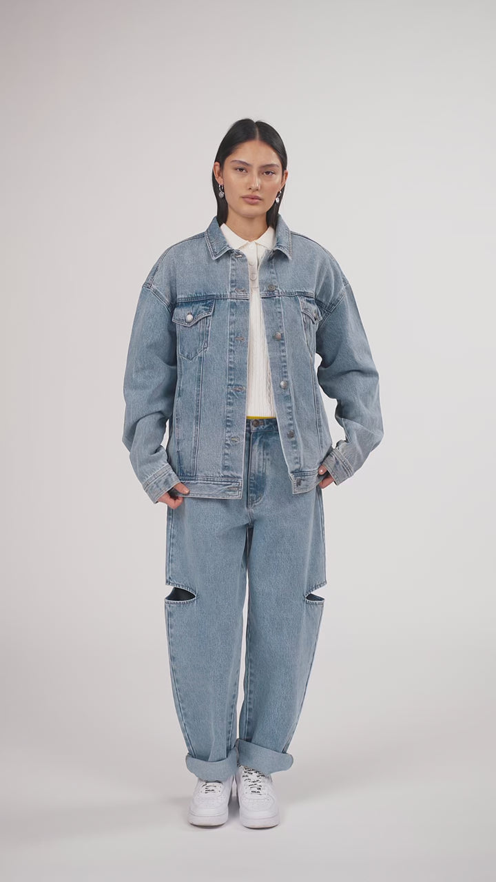 Ein Model in Ganzkörperansicht, das den Look in Bewegung aus verschiedenen Perspektiven zeigt, trägt eine blaue Denimjacke und eine blaue Cut-out-Jeans aus der No Hustle Culture Kollektion, kombiniert mit einem offwhite Strick-Poloshirt, weißen Sneakern und silberfarbenem Schmuck